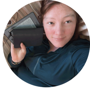 circular cropped photo of white woman holding up a kindle and notebook next to her face.
